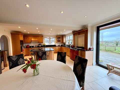 Modern kitchen and dining area with wooden cabinetry, marble countertops, dining table with black chairs, and large glass doors overlooking countryside