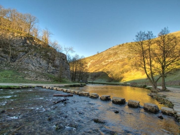 A scenic river valley with golden-hued hills, bare trees, and stepping stones crossing the clear stream under a bright blue sky