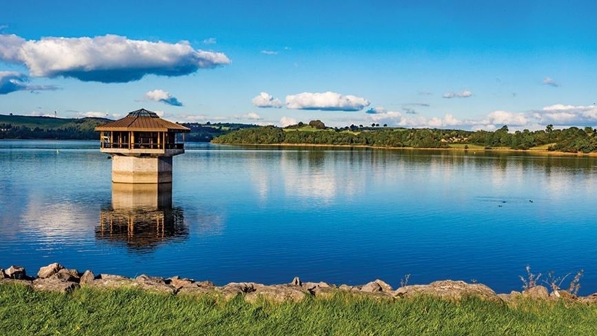 Wooden pump house structure standing alone in calm blue reservoir water with forested shoreline and clear sky