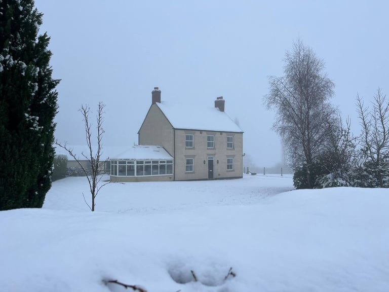 Yellow brick country cottage with chimneys surrounded by snow, bare trees, and a greenhouse in a winter landscape