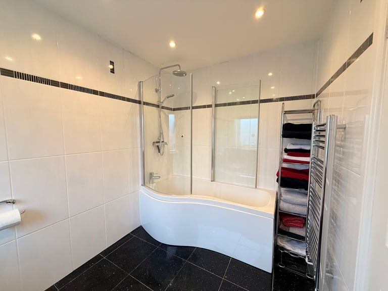 Modern bathroom with curved white bathtub, glass shower enclosure, black floor tiles, white wall tiles, towel rack, and recessed ceiling lights