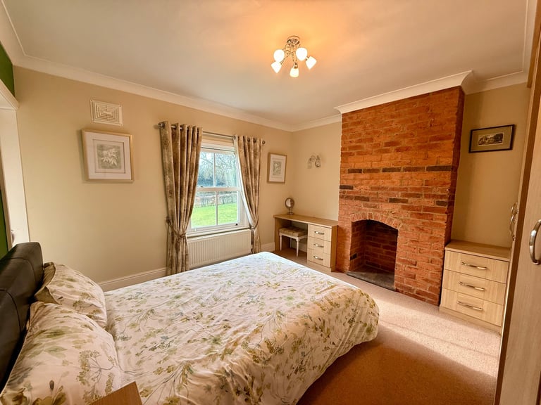Spacious bedroom with double bed, brick fireplace, window with view, cream walls, and traditional furnishings