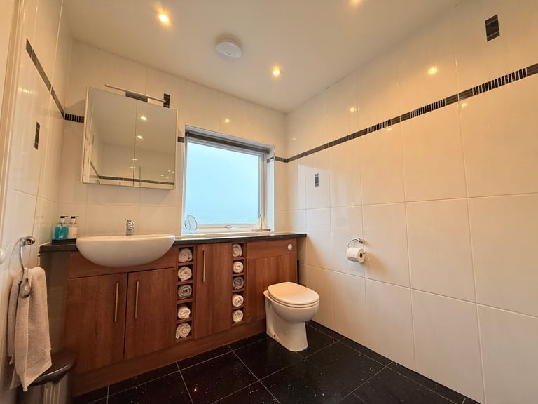 Modern bathroom with wooden vanity, ceramic sink, toilet, white tile walls, dark floor, and window with recessed lighting