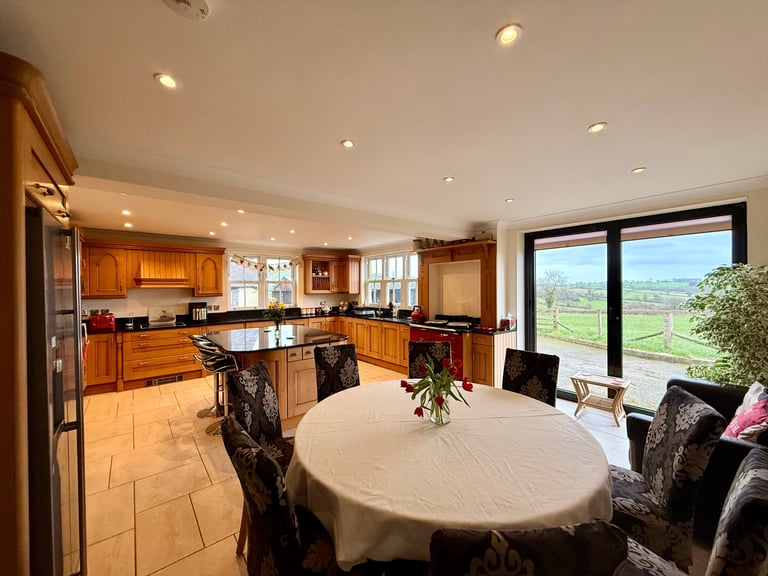 Spacious open-plan kitchen and dining room with wood cabinetry, round white table, and large windows overlooking rural landscape