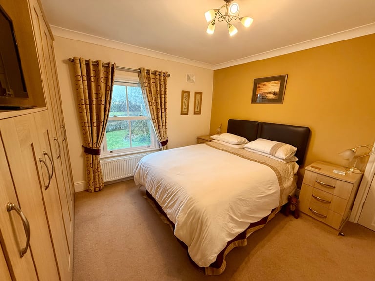 Warm bedroom with yellow walls, double bed with white linens, wooden furniture, window with patterned curtains, and ceiling light fixture